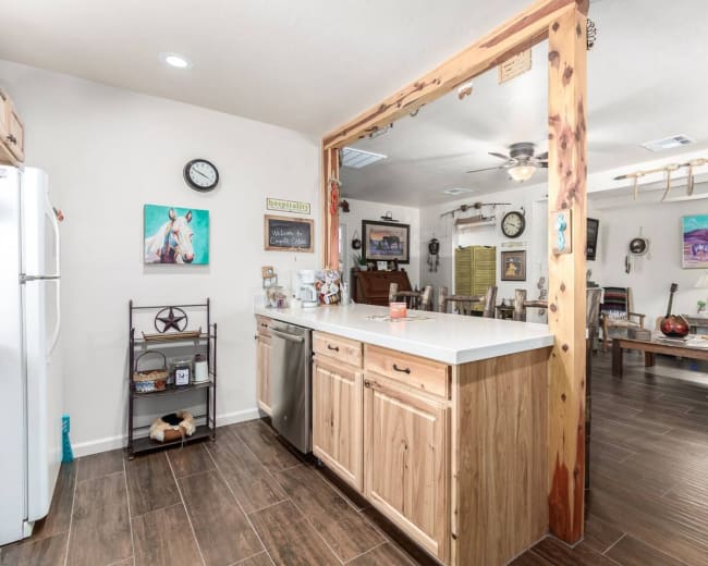 A modern kitchen with wooden cabinetry and a countertop dividing it from a dining area featuring rustic furniture and decorative artwork.