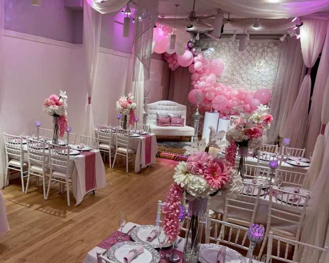 The image shows a decorated event space with white tables, pink floral centerpieces, and a backdrop of pink and white balloons.