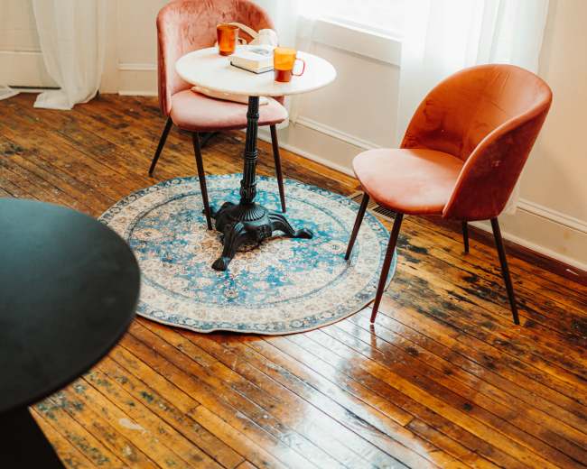 Two pink chairs are positioned around a small table with drinks on it, next to a window with sheer curtains, on a wooden floor with a patterned rug underneath.