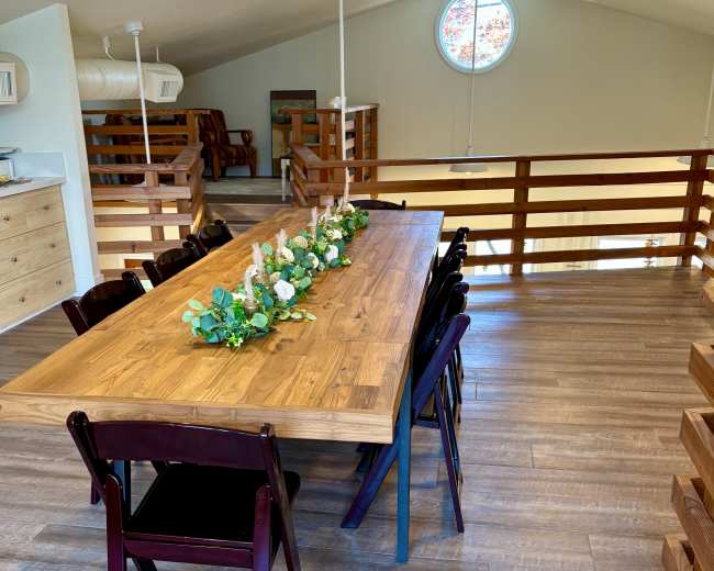 A large wooden dining table is set in a bright, open room with a circular window and a wooden railing.