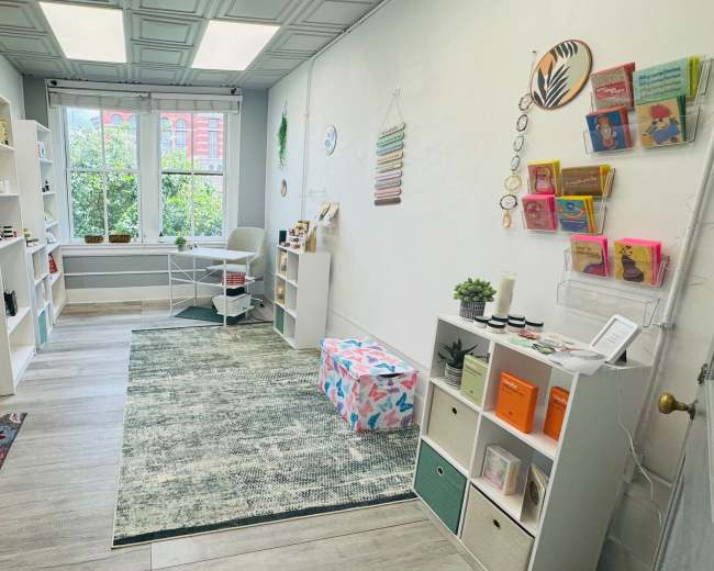 The image shows a bright, organized room with shelves of colorful products, a small seating area, and a green area rug.