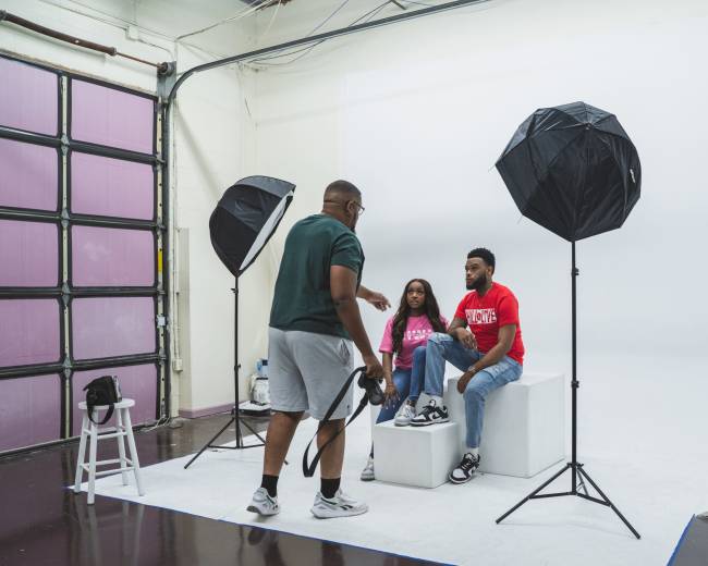 A photographer directs a couple posing on a pedestal in a studio with softbox lights.