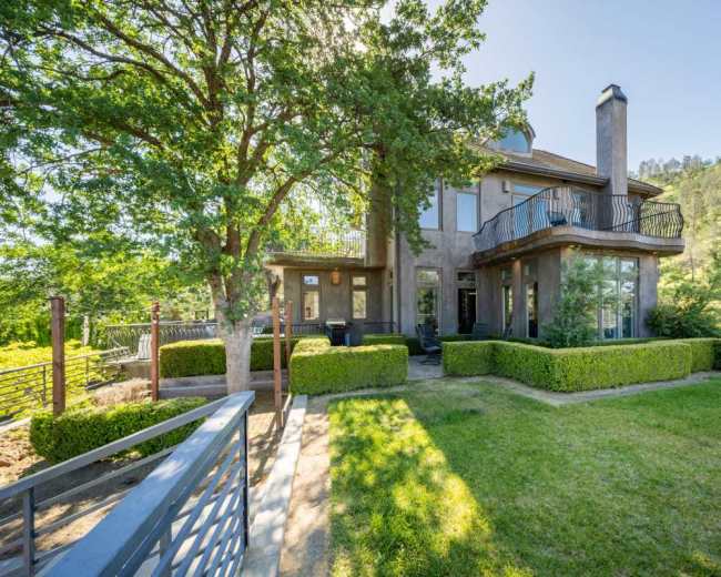 A modern two-story house with large windows and a balcony is surrounded by neatly trimmed hedges and a landscaped yard.