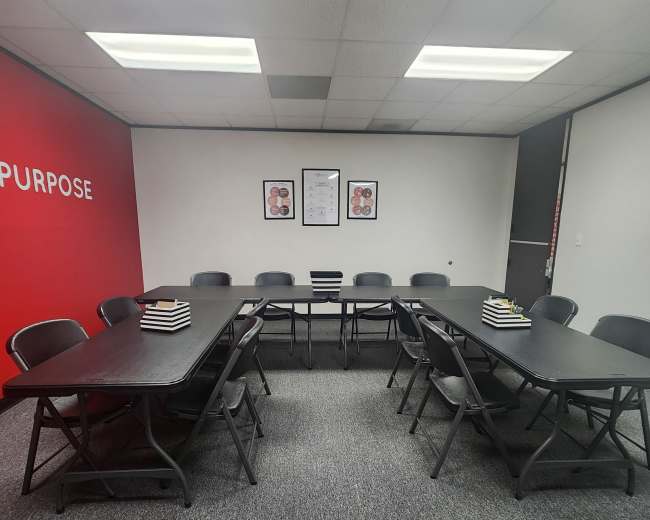 A conference room features a rectangular arrangement of folding tables and chairs, with a red wall displaying the word "PURPOSE" and framed documents on the opposite wall.
