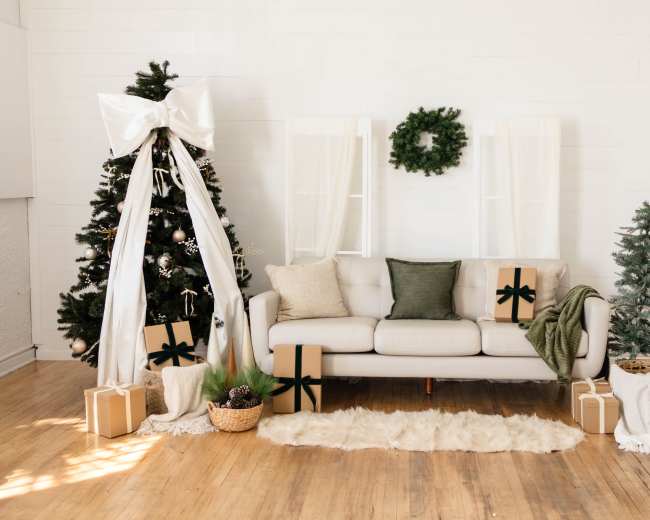 A cozy living room decorated for Christmas features a white sofa flanked by a Christmas tree and a tall arrangement of wrapped presents.
