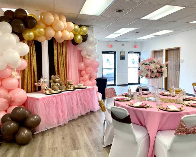 A decorated event space with pink and gold balloons, a table set for a celebration, and floral centerpieces.