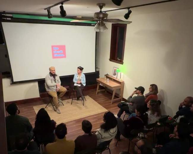 A speaker and a moderator sit on a stage in front of an audience during an event in a small, well-lit room.