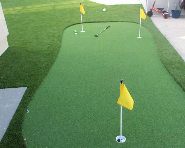A small putting green with three yellow flags and a golf ball sits on a lawn beside a concrete patio.