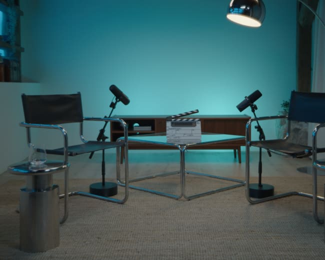 The image shows a minimalist interview setup with two empty chairs, microphones on stands, a coffee table with a clapperboard, and blue lighting in the background.