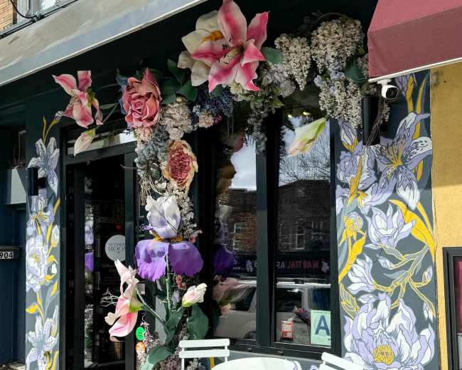 A storefront is adorned with large floral murals and decorations, featuring a small white table and chairs outside.