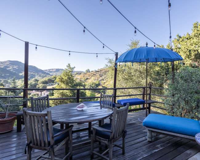 A wooden deck features a round dining table, a lounge chair, and an umbrella, surrounded by trees and hills under a clear sky with string lights overhead.