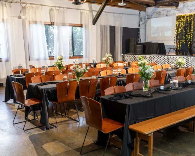 A spacious event venue features several long wooden tables covered with black cloth, each adorned with small flower arrangements, and is set for a gathering.