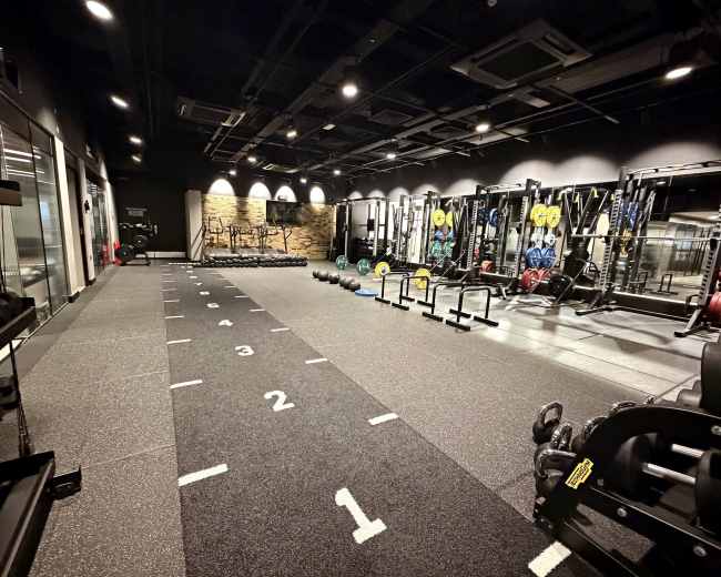 The image shows a modern gym interior with weightlifting equipment, a sprint track marked with numbers, and various exercise tools arranged neatly along the walls.