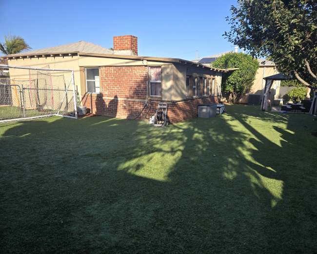 The image shows a backyard with artificial turf, a brick house, a chimney, and a goalpost in the corner.