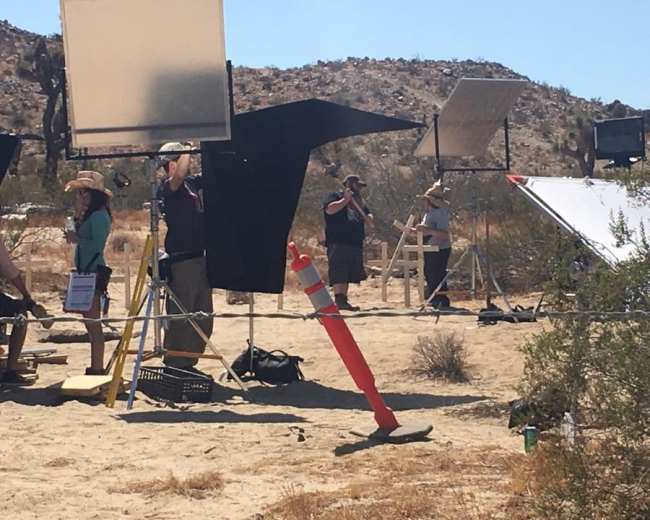 A film crew is set up in a desert landscape with various lighting equipment and a wooden structure in the background.
