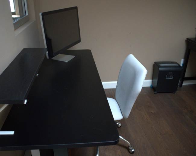 The image shows a simple black desk with a raised shelf, a computer monitor, and an empty white chair in a room with brown walls and wooden flooring.