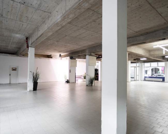 A spacious, empty indoor area with concrete walls and columns, featuring tiled flooring and minimal furnishings.