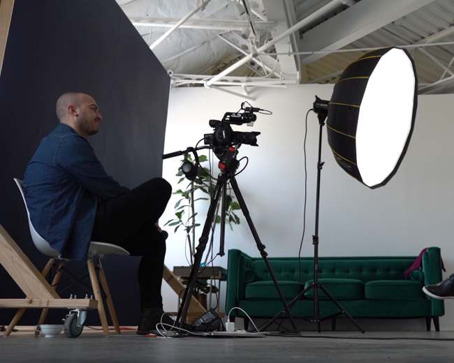 A man and a woman are seated in a well-lit studio, with a camera on a tripod focused on the woman, who is speaking while the man listens.