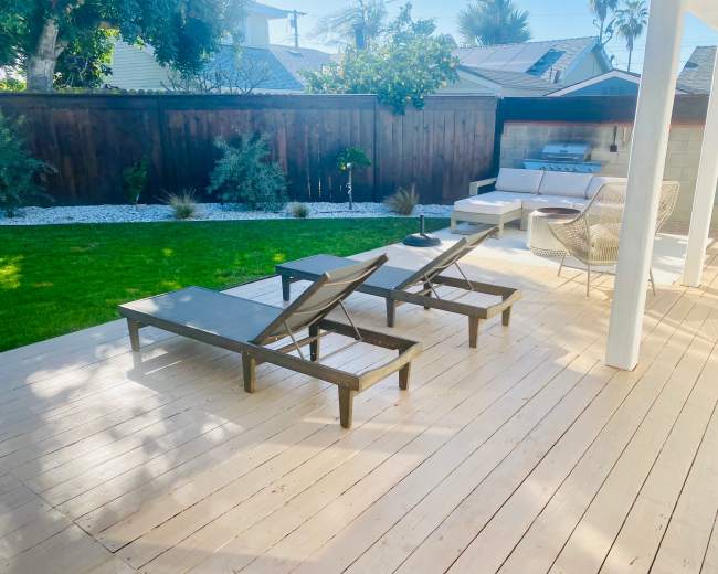 The image shows a sunny patio with two lounge chairs and a small table overlooking a landscaped backyard.