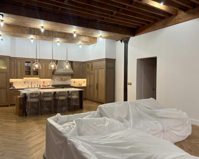The image shows a partially furnished room featuring two large, white, covered furniture pieces, a kitchen area with wooden cabinetry and pendant lighting, and exposed wooden beams on the ceiling.