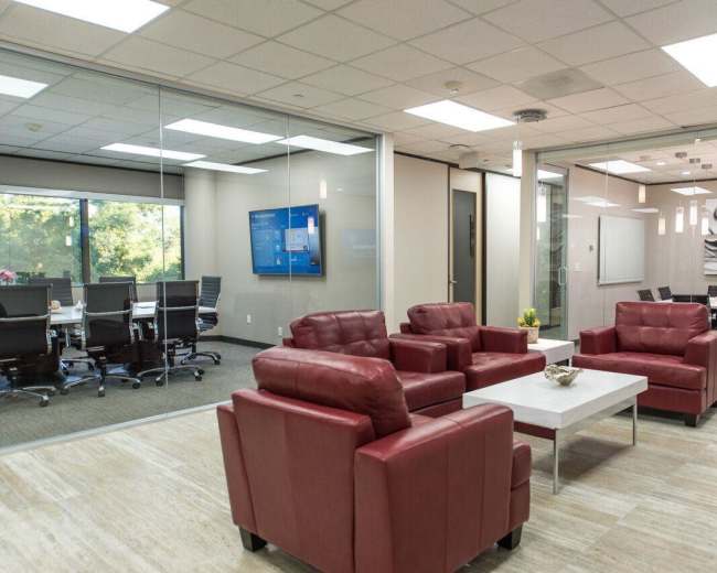 A modern office space featuring red leather chairs, a coffee table, and glass-enclosed conference rooms with a view of greenery outside.