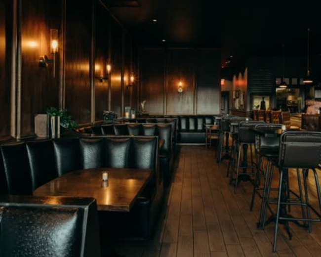 The image shows a dimly lit restaurant interior with dark wooden walls, leather seating, and a bar area in the background.
