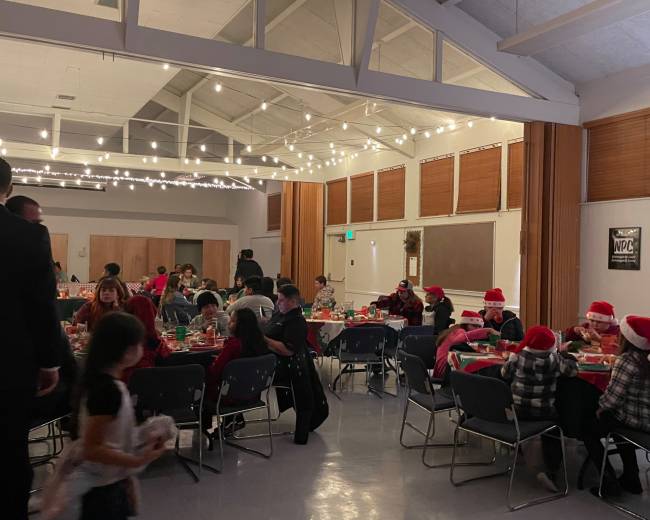 A group of people wearing Christmas hats gathers around tables decorated for a holiday party, with string lights illuminating the room.