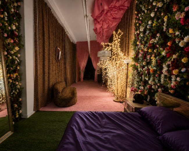 The image shows a decorated interior space resembling a whimsical bedroom with floral walls, soft lighting, and a pink carpet.