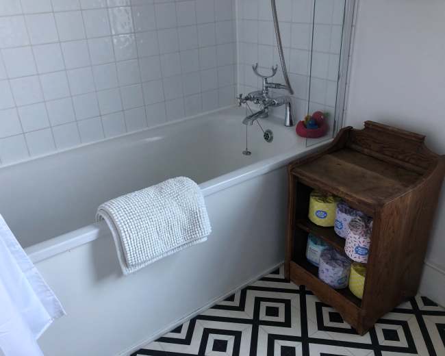 The image shows a bathroom with a white bathtub, a wooden shelf holding rolled towels, and a tiled floor featuring a black and white geometric pattern.