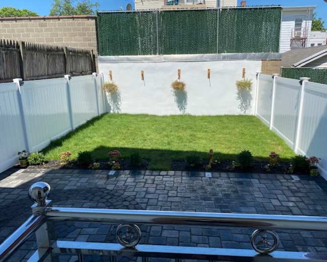 The image shows a fenced backyard with neatly trimmed grass and small flower beds along the edges, featuring white fencing and a clean stone patio.