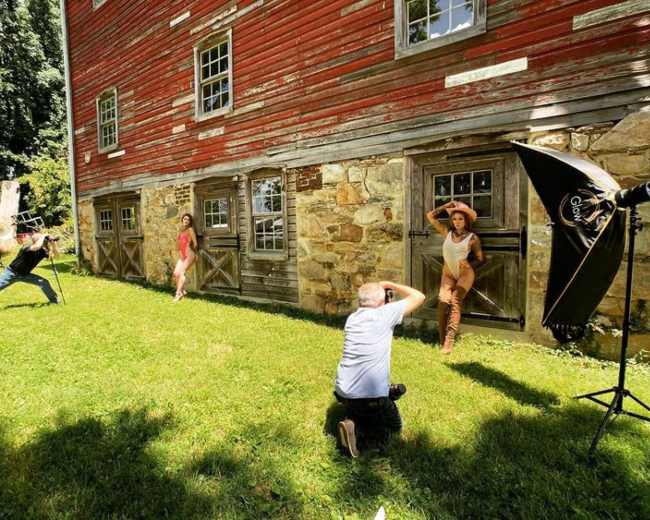 A photographer captures models posing in front of a rustic barn while an assistant sets up lighting equipment.