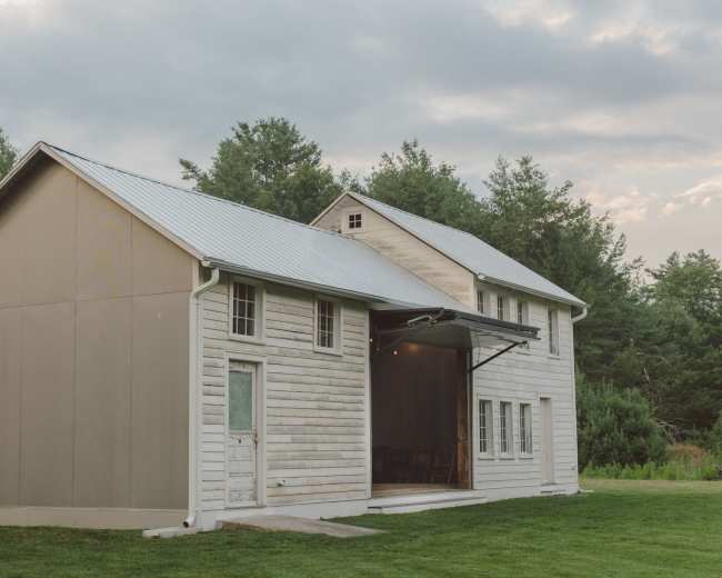 A two-story, modern farmhouse with a metal roof, situated on a grassy area surrounded by trees.