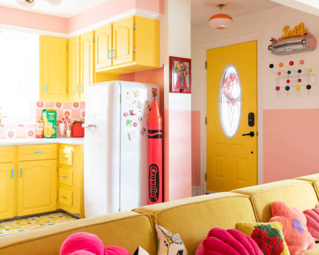 A brightly colored kitchen features yellow cabinets, a white refrigerator adorned with magnets, and a large red crayon decoration alongside a cheerful yellow door.