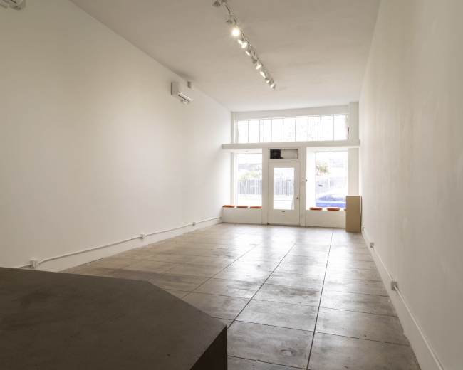 The image shows a spacious, empty interior of a room with white walls, a tiled floor, and large front windows.