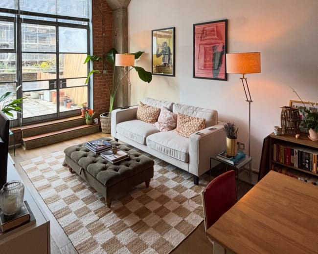 The image depicts a modern living room with a white sofa, a coffee table, a dining area, and large windows allowing natural light to enter.