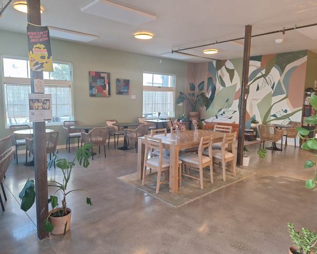 The image shows a spacious café with wooden tables surrounded by plants and a colorful mural on the wall.
