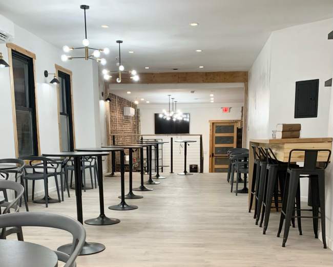 A sparsely-furnished interior of a restaurant or café with rows of black tables and chairs along a light-colored wooden floor.