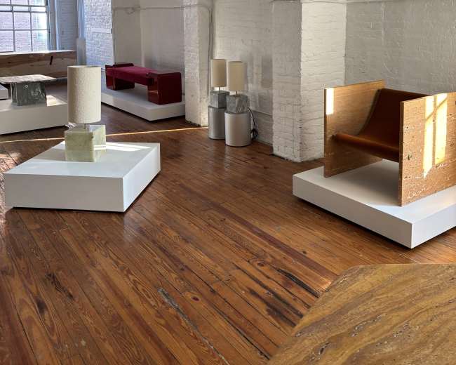 The image shows an art gallery with wooden floors and white walls, featuring various modern furniture pieces displayed on minimalist stands.