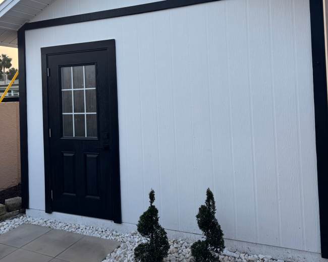 A white shed with a black door and two small, neatly trimmed shrubbery plants is positioned in a paved area.