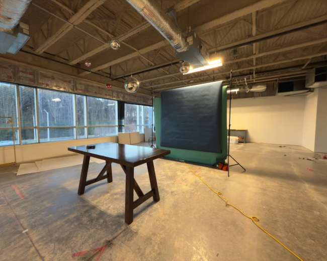 The image shows an empty room with a large window, a wooden table in the center, and a backdrop setup in the corner.