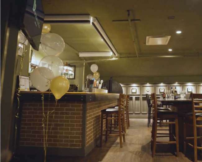 The image shows a spacious bar area with wooden tables and chairs, a brick counter, and party balloons in the corner.