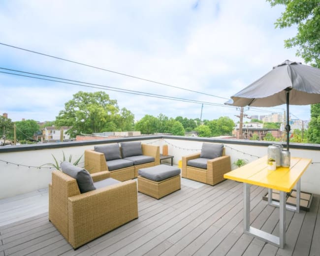 The scene features a rooftop terrace with wicker seating, a yellow table, and an umbrella, surrounded by greenery and city buildings in the background.