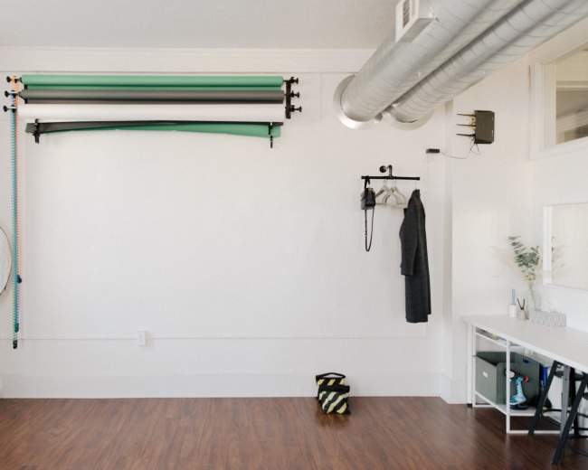 A minimalist studio space with a white wall, a clothing rack, a reflector on a stand, and a neatly organized tabletop beneath a metal ventilation duct.