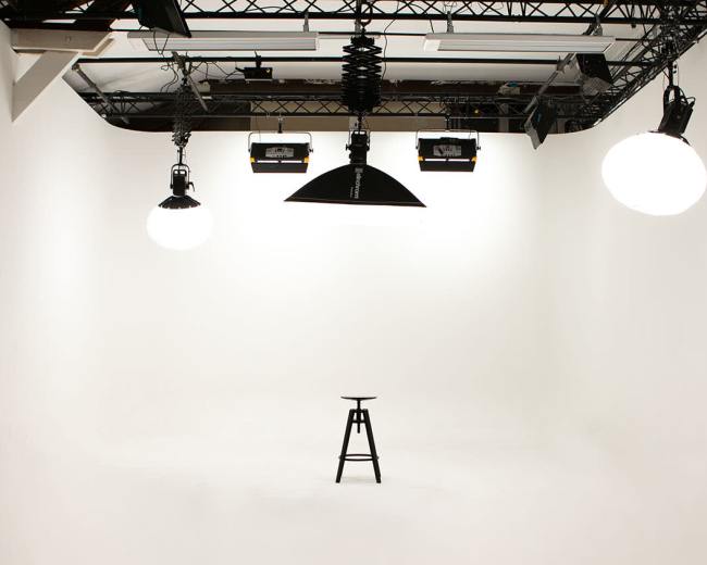 A minimalistic photography studio features a single black stool illuminated by overhead lights in a white room.