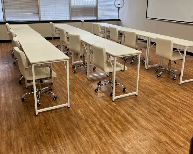 The image shows a classroom with several white tables and chairs arranged in rows, along with a large projection screen at the front.