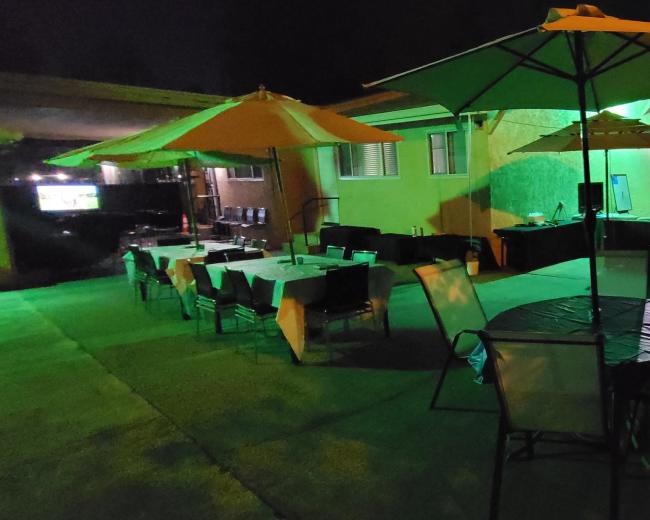 A nighttime outdoor gathering features tables and chairs under yellow umbrellas, with green lighting illuminating the area.