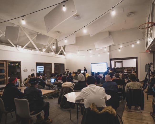 A group of people is seated in a large room with a presentation screen at the front, under hanging lights and basketball hoop.