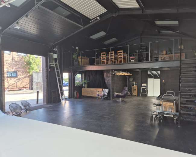The image shows a spacious industrial-style interior with a black ceiling and walls, featuring an open area on the ground floor and a mezzanine level with chairs and plants.
