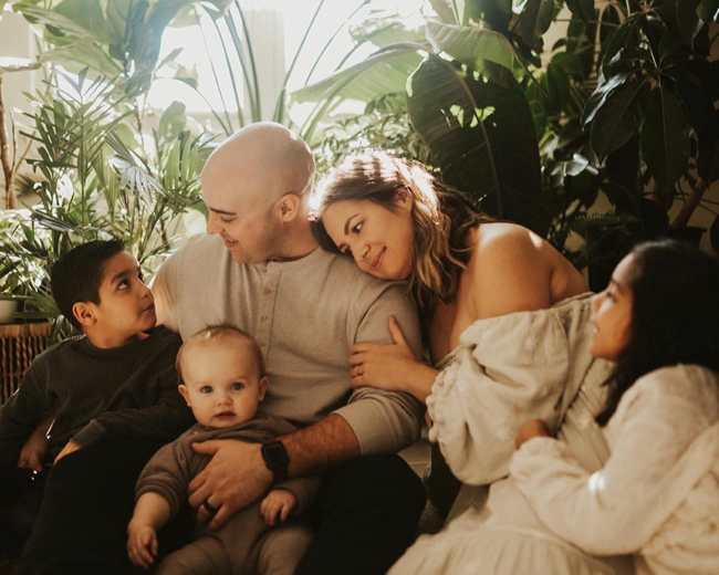 A family sits together on the floor surrounded by various indoor plants, with two children looking at their parents and a baby in the mother's lap.