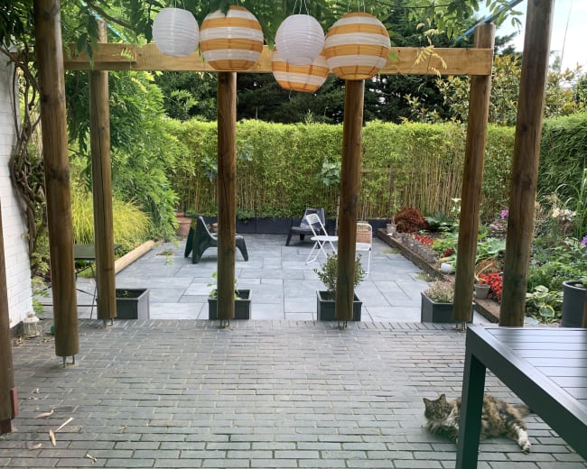 The image shows a patio area with wooden beams and lanterns, overlooking a landscaped garden with seating and a cat lying on the ground.
